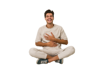 Young caucasian man sitting on the floor isolated on white background laughs happily and has fun keeping hands on stomach.