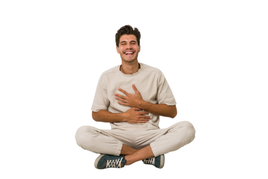 Young caucasian man sitting on the floor isolated on white background laughs happily and has fun keeping hands on stomach.