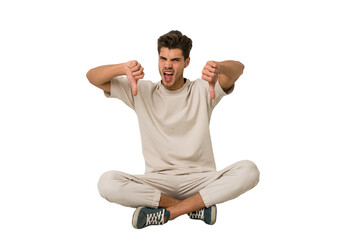 Young caucasian man sitting on the floor isolated on white background showing thumb down and...