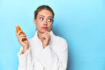 Blonde in bathrobe with sunscreen cream in studio looking sideways with doubtful and skeptical...