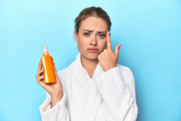 Blonde in bathrobe with sunscreen cream in studio showing a disappointment gesture with forefinger.