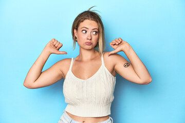 Blonde young woman in white top on blue background feels proud and self confident, example to...
