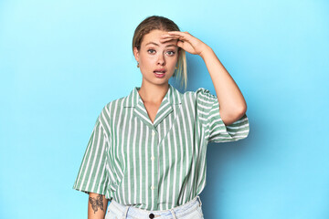 Blonde young woman in green striped shirt on blue background shouts loud, keeps eyes opened and...