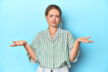 Blonde young woman in green striped shirt on blue background doubting and shrugging shoulders in questioning gesture.