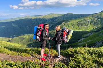 Two hikers, man and woman with backpacks and sleeping pads, stand facing each other on mountain...