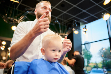 Young child getting haircut in modern barbershop. Hairdresser making final touches in new haircut, shaping child's hair by hands.