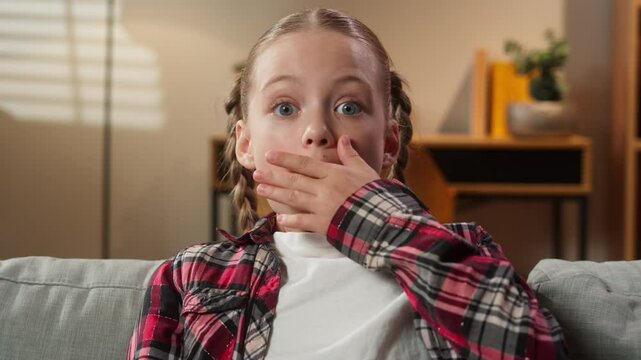 Portrait surprised little school girl child kid daughter preteen looking at camera at home sofa couch big eyes close mouth with hand feel stunned amazed unexpected news shocked unbelievable surprise