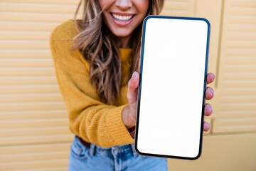 A close-up shot of a cheerful woman in a yellow sweater and blue jeans holding a large smartphone with a blank white screen in front of her. 