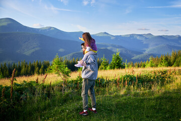 Fototapeta na wymiar Father carries his young daughter on his shoulders, both enjoying scenic mountain view. They surrounded by lush greenery and rolling hills under clear blue sky.
