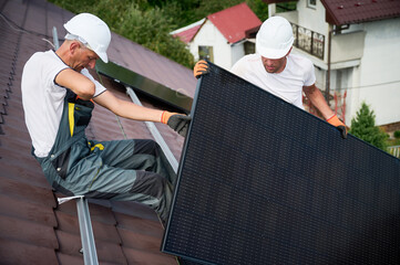 Workers building solar panel system on rooftop of house. Two men installers in helmets installing photovoltaic solar module outdoors. Alternative, green and renewable energy generation concept.
