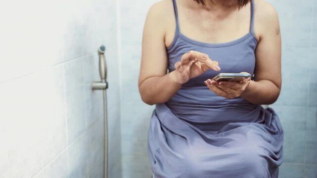 asian woman having trouble passing stool while using her phone on the toilet, struggling with constipation and showing nomophobia behavior from constant mobile phone use