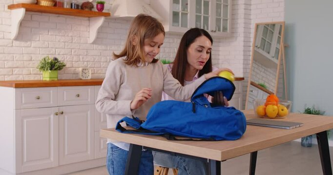 Caring mother helping son to pack schoolbag, putting tablet and snack, wishing good day at school, posing at kitchen. Schoolboy preparing for school. Family, parenting and studying concept