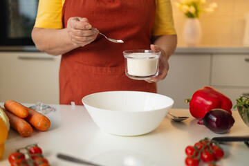 A senior woman in a yellow shirt and brown apron adds sugar to a bowl while preparing a meal in a...