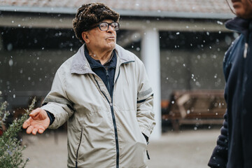 Elderly man wearing winter clothing stands outdoors during snowfall, observed near a weathered...