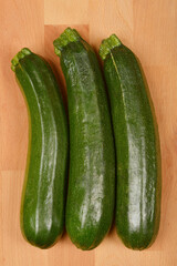 Three green zucchinis are arranged vertically on a light brown wooden surface