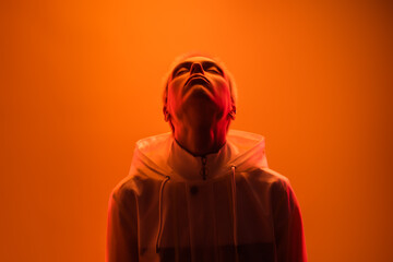 A person wearing a hoodie looks upwards, backlit in warm orange lighting. The image conveys a sense...