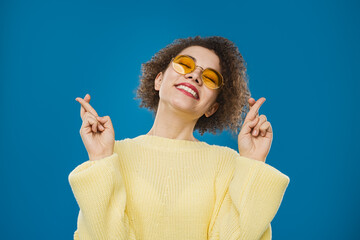 A smiling woman wearing a yellow sweater and sunglasses crosses her fingers, symbolizing hope and...