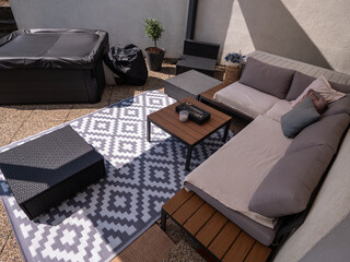 Fototapeta premium Modern aluminum patio with outdoor sofa, hot tub, coffee tables, and decorative rug on stone tiles, partially shaded, with potted plant and wall in the background.