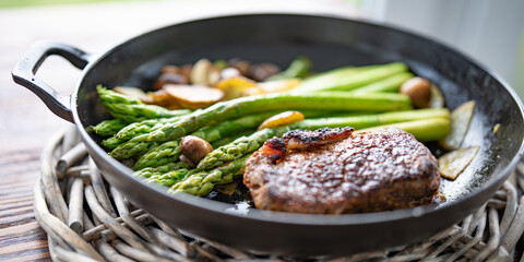 Crispy roasted beef steak with green asparagus, mushrooms, and potato slices in a cast iron pan. Close-up with shallow depth of field.