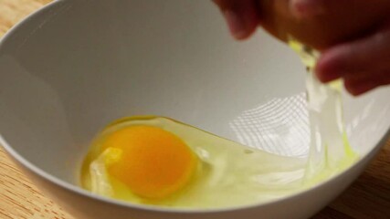 An egg is being cracked and poured into a white bowl, handheld, slow motion