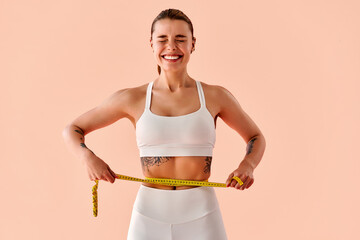 Smiling fit woman in white activewear measuring her waist with a tape measure on soft peach background. Concept of fitness progress, motivation, and healthy lifestyle.