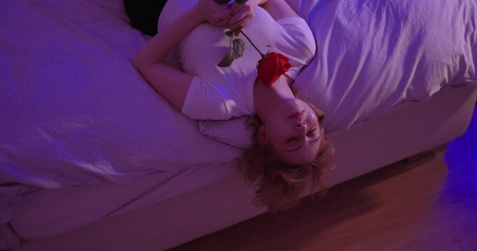 young blonde woman lies in bed and lets her head hang over the edge of the bed and looks at rose