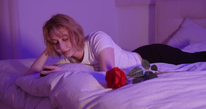young blonde woman lying on bed and scrolling through smartphone next to her a rose