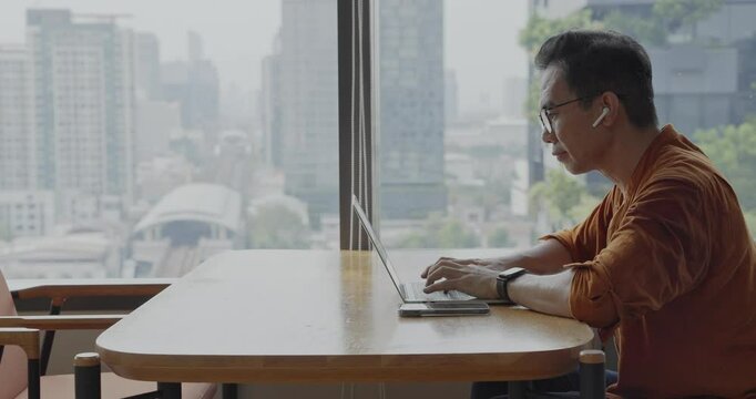 Senior Asian businessman wearing Bluetooth headset celebrates success during online meeting in modern co-working space. Remote work and business communication concept.
