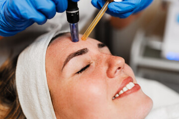 Beautician wearing blue gloves uses a microneedling pen to apply serum to forehead of client with...