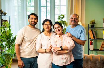 Smiling at camera, Indian family of four standing together in cozy and modern living room