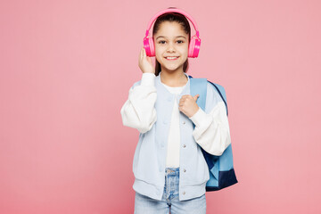 Young school girl 8-9 years old she wear blue casual clothes backpack listen to music in headphones show thumb up isolated on plain pink background children portrait. Childhood kids education concept.