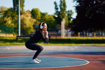 Young sportswoman doing squats and listening music with headphones in a basketball court