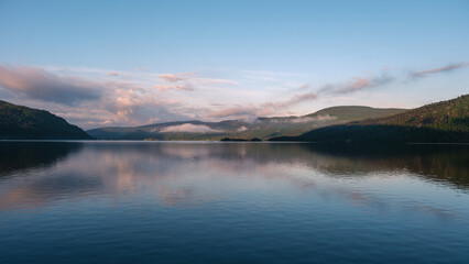 Fototapeta premium Morning light casts a peaceful glow over the tranquil waters of a lake in Norway. Mist gently rises from the surrounding hills, creating a serene atmosphere perfect for relaxation and inspiration.