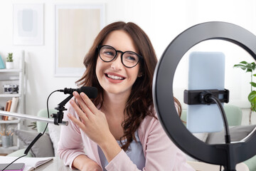 Smiling young female content creator and influencer at home, using a smartphone and ring light,...