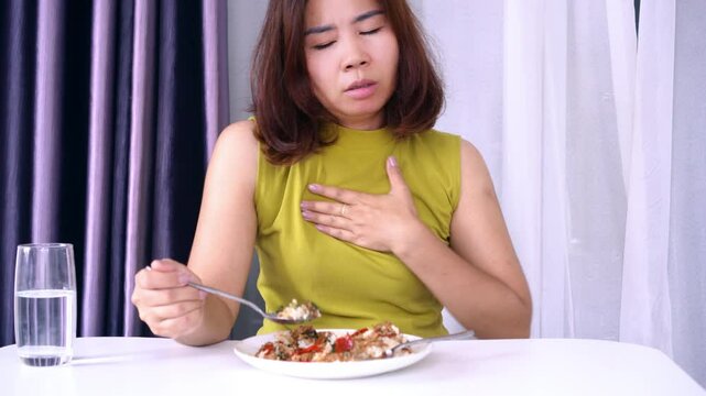 Asian woman suffering from Gastroesophageal Reflux Disease or GERD holding her sore throat in discomfort from symptoms like heartburn and acid reflux after eating spicy food