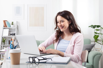 Happy young woman at home, sitting at a cozy desk and smiling at her laptop. Representing a positive remote work or online learning, video call, or digital collaboration in comfortable home setting
