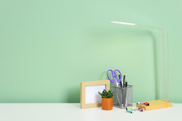 Stylish workplace with lamp, stationery and plant on white desk near light green wall. Space for text