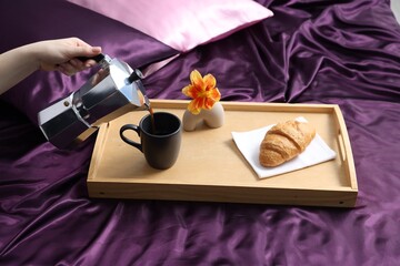 Woman pouring coffee from moka pot into cup on bed with purple silk linens, closeup