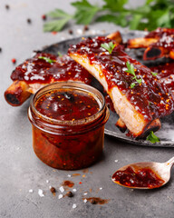 A jar of rich red chili sauce placed beside glazed grilled pork ribs garnished with parsley on a dark grey plate, with sauce dripping off a spoon.