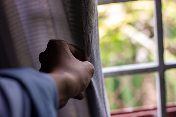 Close-up of a hand pulling back a grey curtain to look out a window with a blurry view of green foliage. Represents home, morning, and observation.

