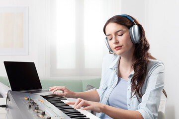 Young woman at home, playing a music keyboard and listening through headphones. She is learning and...