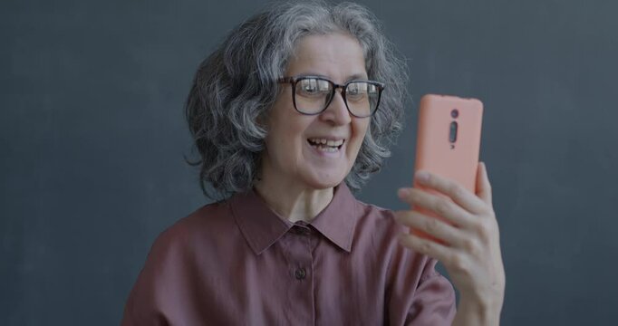 Senior lady making online video call with smartphone and earphones talking waving hand on gray color background. People and communication concept.