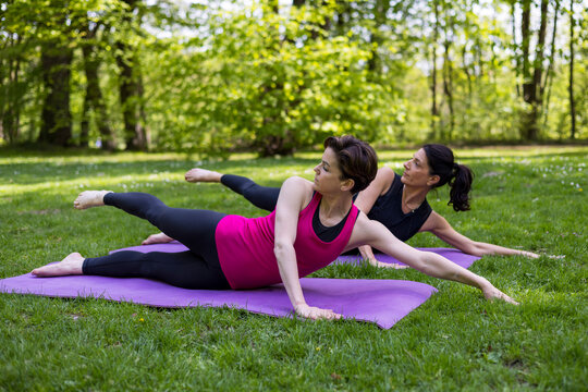 Two fit women performing side leg lifts and extensions on purple mats during a outdoor pilates session in a lush green park, focusing on core strength, flexibility, wellness, and active lifestyle.