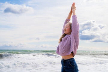 Young woman stretching arms up on beach with ocean waves under blue sky, morning practice, eyes...