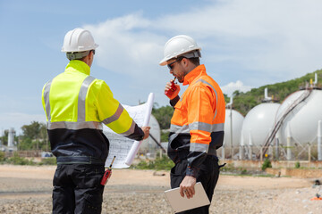 Two engineers wearing safety uniforms and helmets review construction blueprints and discuss an...