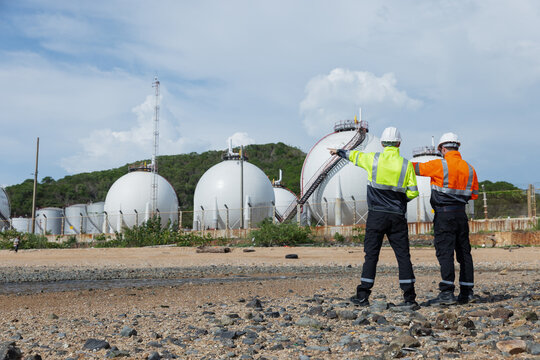 Two environmental engineers wearing safety gear observe and discuss an industrial facility with large spherical storage tanks, likely evaluating environmental safety or maintenance conditions.
