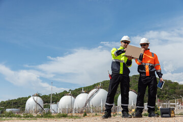 Two engineers wearing high-visibility safety gear use a laptop and tablet for environmental monitoring and industrial inspection near spherical storage tanks in an outdoor coastal facility.