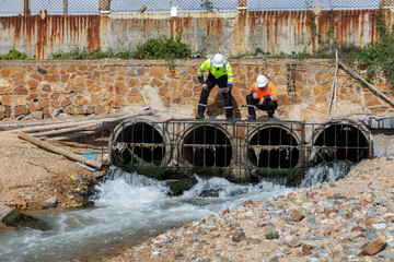 Two environmental engineers inspect industrial drainage pipes with flowing wastewater, assessing...