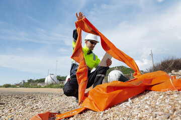 An environmental engineer wearing safety gear inspects coastal waste, holding an orange tarp and using a digital tablet to record data 