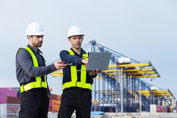 Two port workers wearing reflective safety vests and helmets analyze operations on a laptop at a shipping yard, with large yellow cranes and cargo containers in the background.